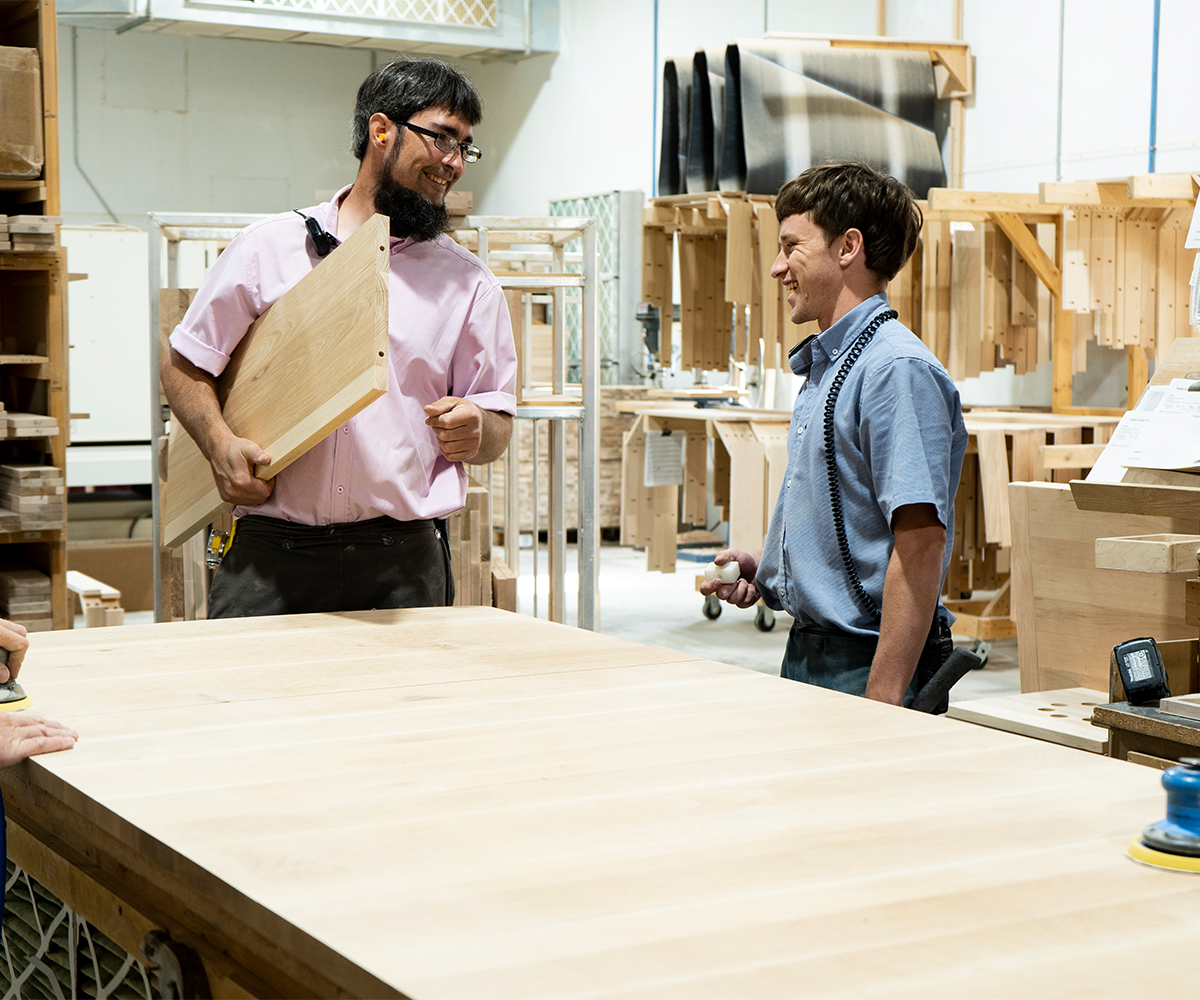 Two woodshop craftsmen review a wood panel during order handoff.