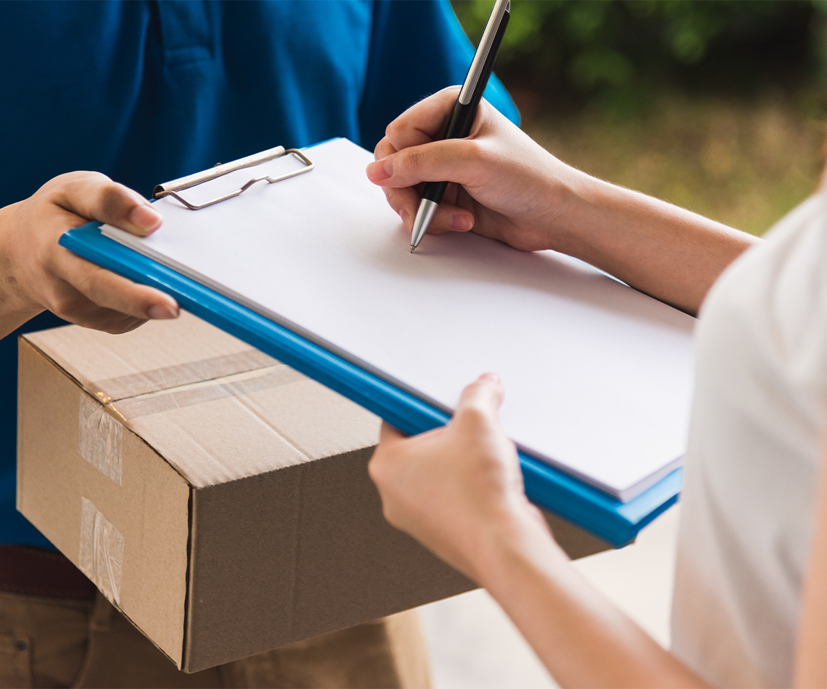 Customer signs delivery paperwork on clipboard beside a shipping box.